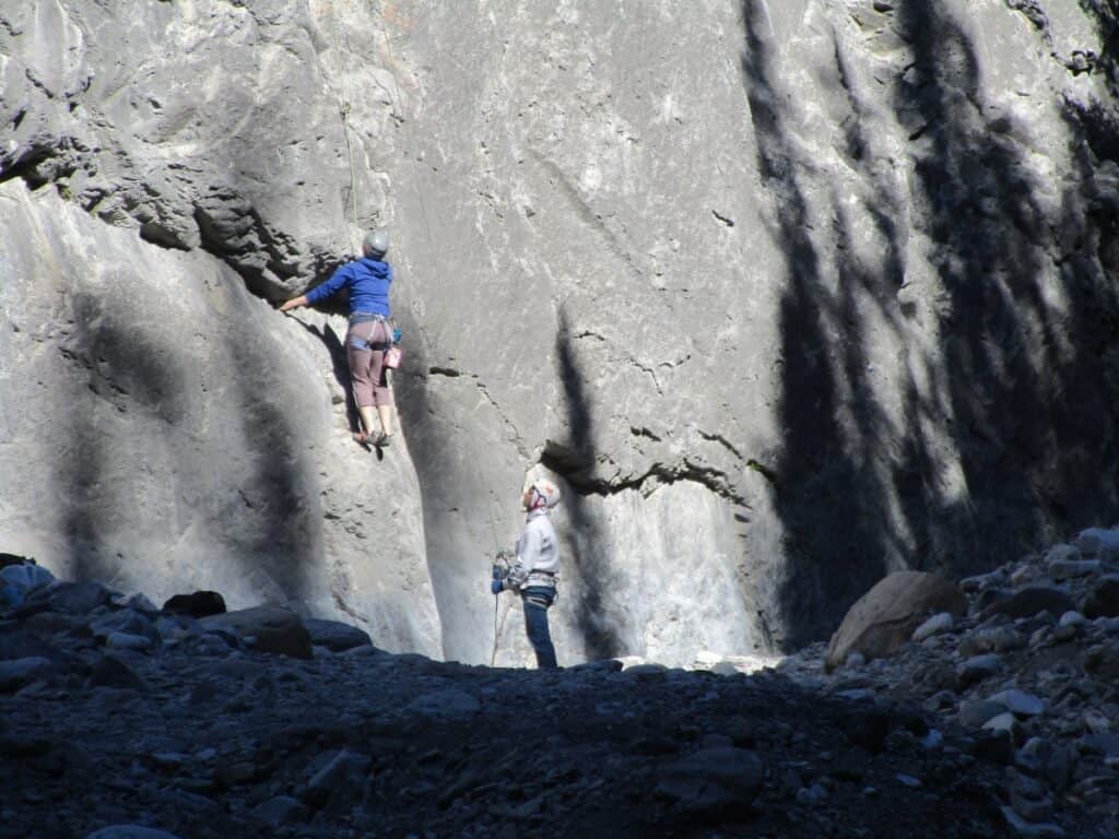A climber in blue scaling a sheer rock face in Grotto Canyon near Canmore, with a partner on the ground securing the rope, amidst the shadows and rugged terrain.