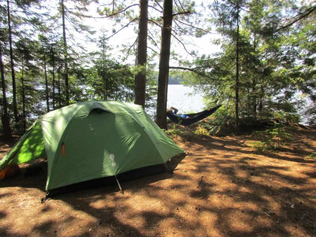 A tranquil campsite nestled among tall pine trees on the edge of Lake Opeongo in Algonquin Provincial Park, featuring a green tent in the foreground and a person relaxing in a hammock in the background. The campsite, dappled with sunlight filtering through the trees, offers a serene view of the lake.