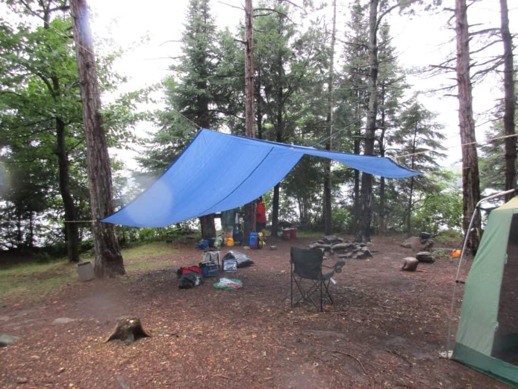 A campsite in Algonquin Provincial Park is set up with a blue rain tarp shelter strung between trees, overlooking Lake Opeongo. Underneath the tarp are camping chairs and gear, suggesting a cozy gathering spot for campers. The forest floor is damp, indicating recent rain, and the backdrop is a dense arrangement of coniferous trees, enhancing the secluded and rustic ambiance of the campsite.