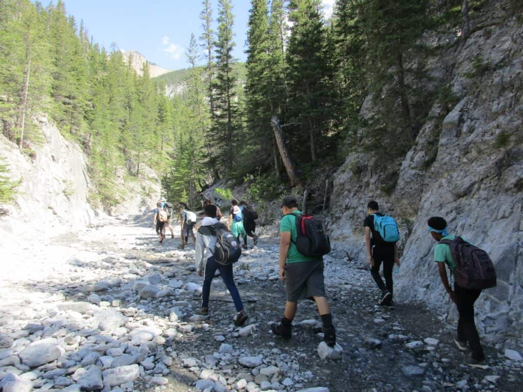 A group of young hikers with backpacks navigating a rocky section of the Grotto Canyon Trail near Canmore, surrounded by steep canyon walls and tall green trees.