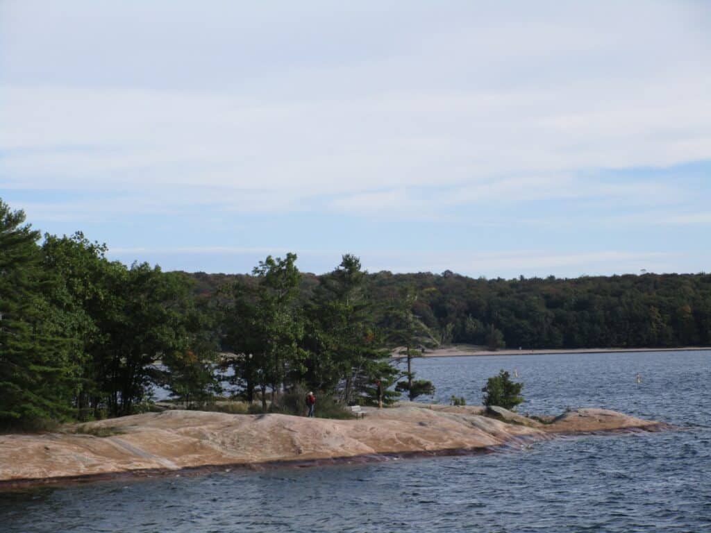 Scenic view of the Twin Points Trail in Killbear Provincial Park, featuring a granite shoreline leading into a serene lake, with a backdrop of dense greenery and a clear sky with wispy clouds.