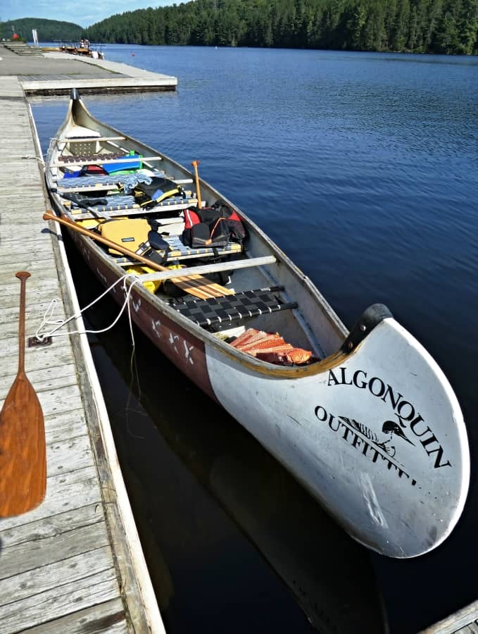 A fully loaded voyageur canoe from Algonquin Outfitters is docked at a wooden pier on the calm waters of Lake Opeongo in Algonquin Park. The canoe is packed with various outdoor gear, paddles, and life jackets, ready for an adventure in the wilderness under a clear blue sky.