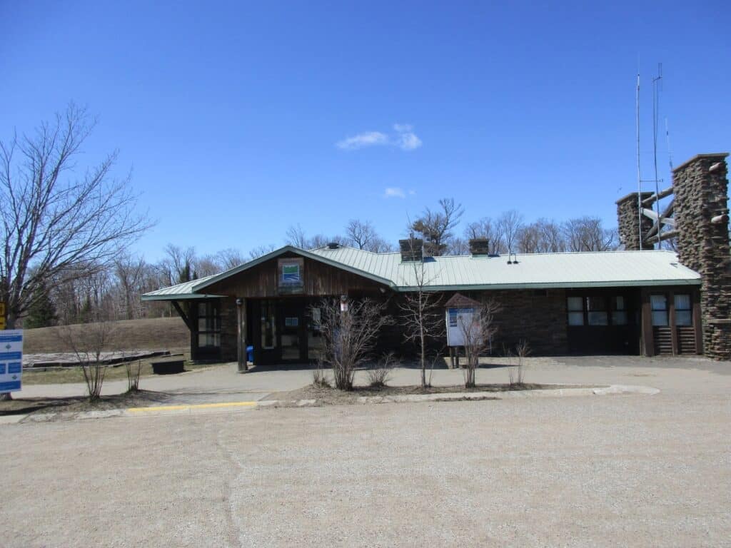The West Gate of Algonquin Provincial Park is depicted on a clear day, featuring a rustic building with stone pillars and informational signage. The area is barren of visitors, with leafless trees suggesting early spring or late fall, and the blue sky offers a sharp contrast to the earthy tones of the park’s entrance structure.
