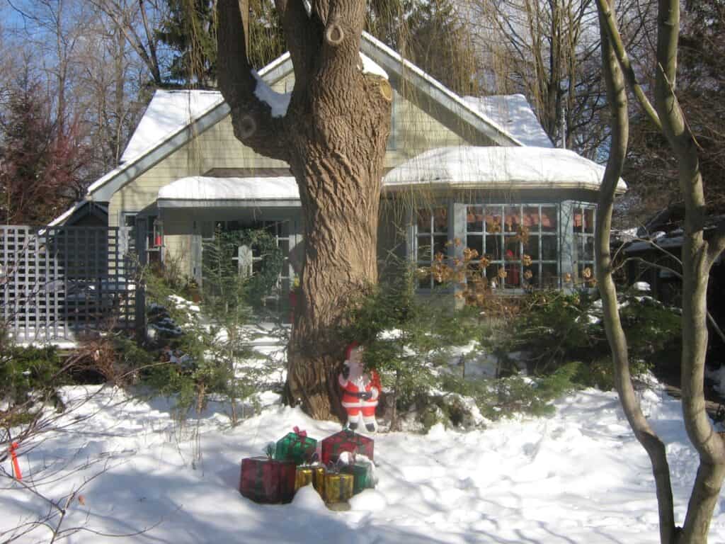 A cozy Algonquin Island home on the Toronto Islands covered in a blanket of snow, featuring a large tree in the front yard with Christmas decorations and a snowman, and a sunroom decorated with festive ornaments visible through the windows.