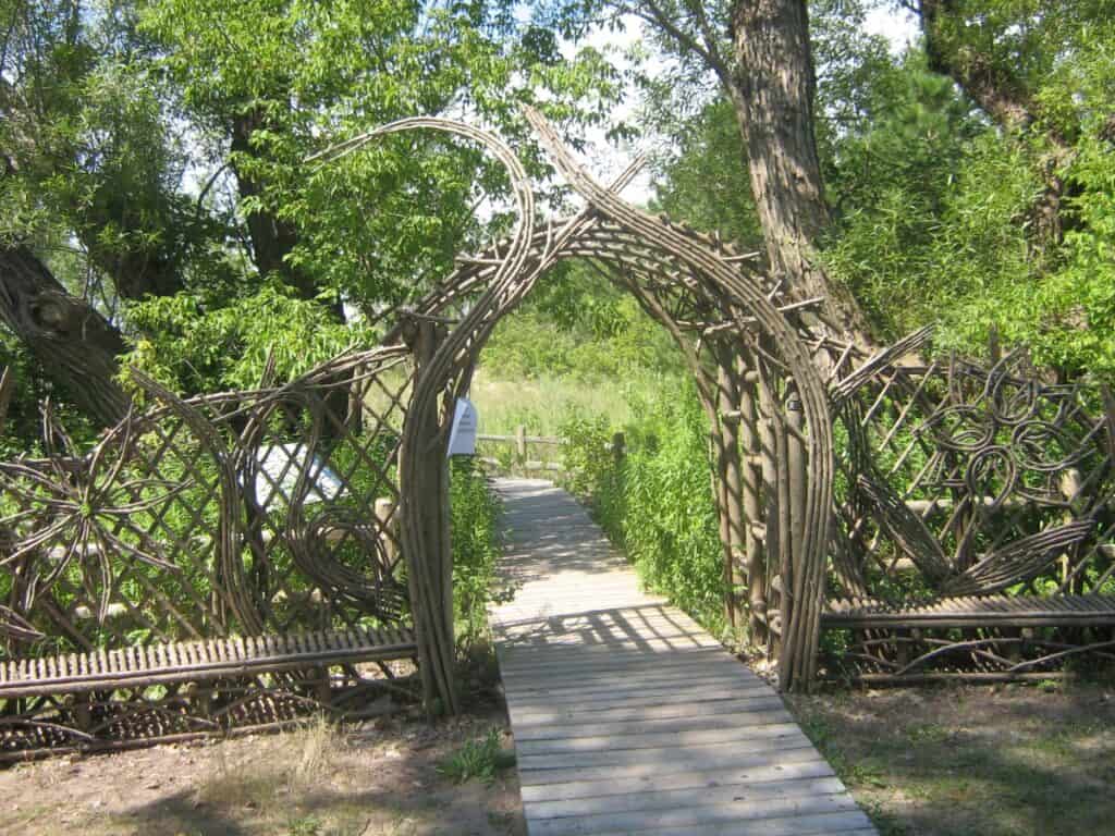 Rustic archway entrance to Franklin Garden on the Toronto Islands, crafted from intertwining branches and bicycle wheel rims, leading down a wooden boardwalk flanked by lush greenery