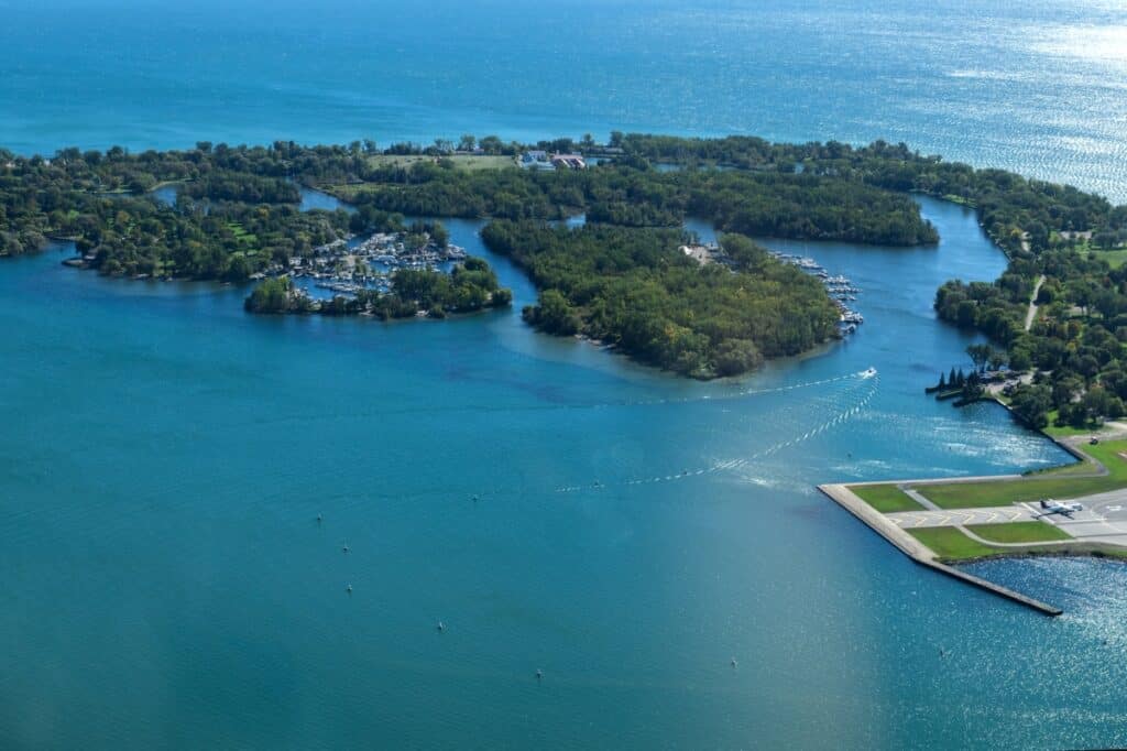 Visiting the Toronto Islands with an aerial view, showcasing the intricate waterways, marinas, and green spaces, set against the expansive blue of Lake Ontario, with boats leaving gentle wakes on the water.