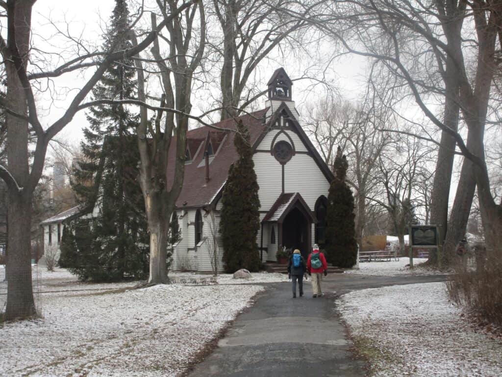 Two people walking towards St. Andrews by the Lake Church on the Toronto Islands, a quaint white church with a prominent belfry, surrounded by leafless trees and a dusting of snow on the ground, conveying a peaceful winter scene.