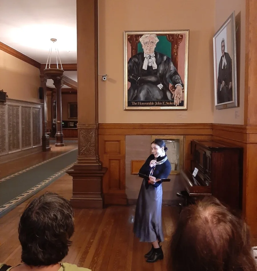 An actor, portraying an artist, stands animatedly speaking in a corridor of the Ontario Legislative Building, in front of a portrait labeled 'The Honourable John S. MacDonald' while listeners are seated and attentively engaged