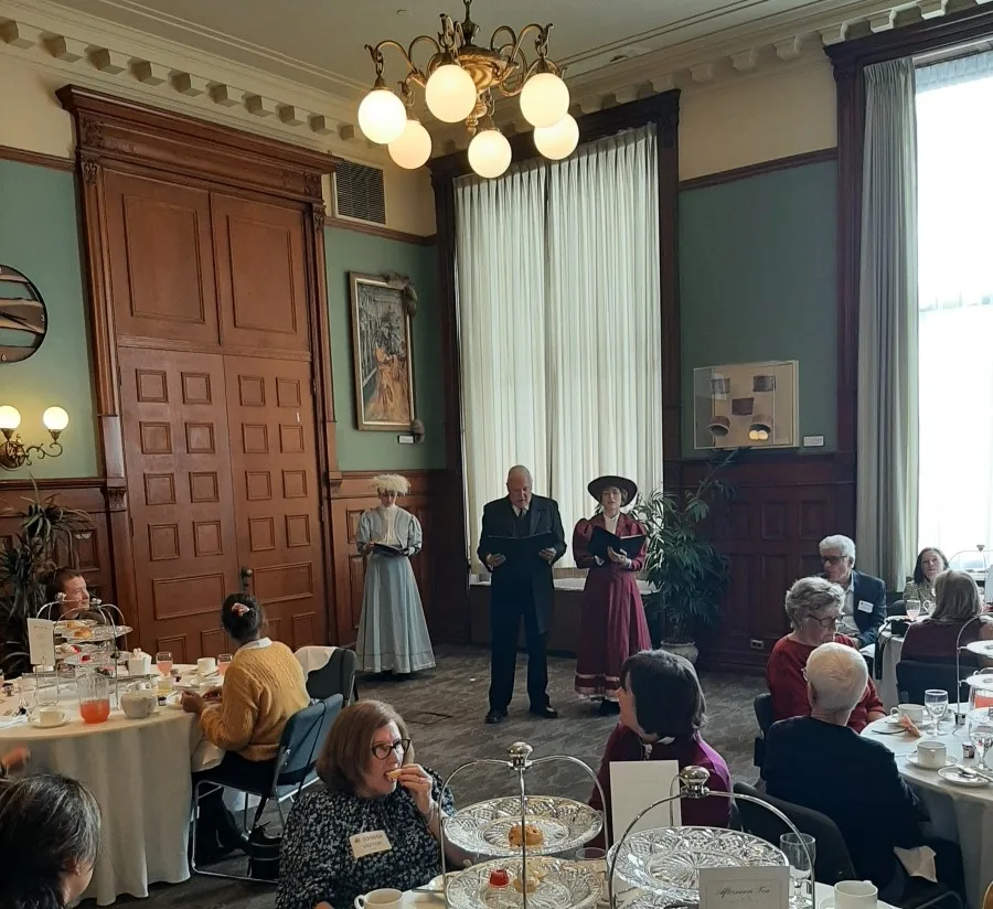 Actors in period costumes perform at an afternoon tea service during a tour of the Ontario Legislative Building, standing elegantly against the backdrop of a stately room with high ceilings, chandeliers, and large windows, while guests are seated at tables enjoying the event