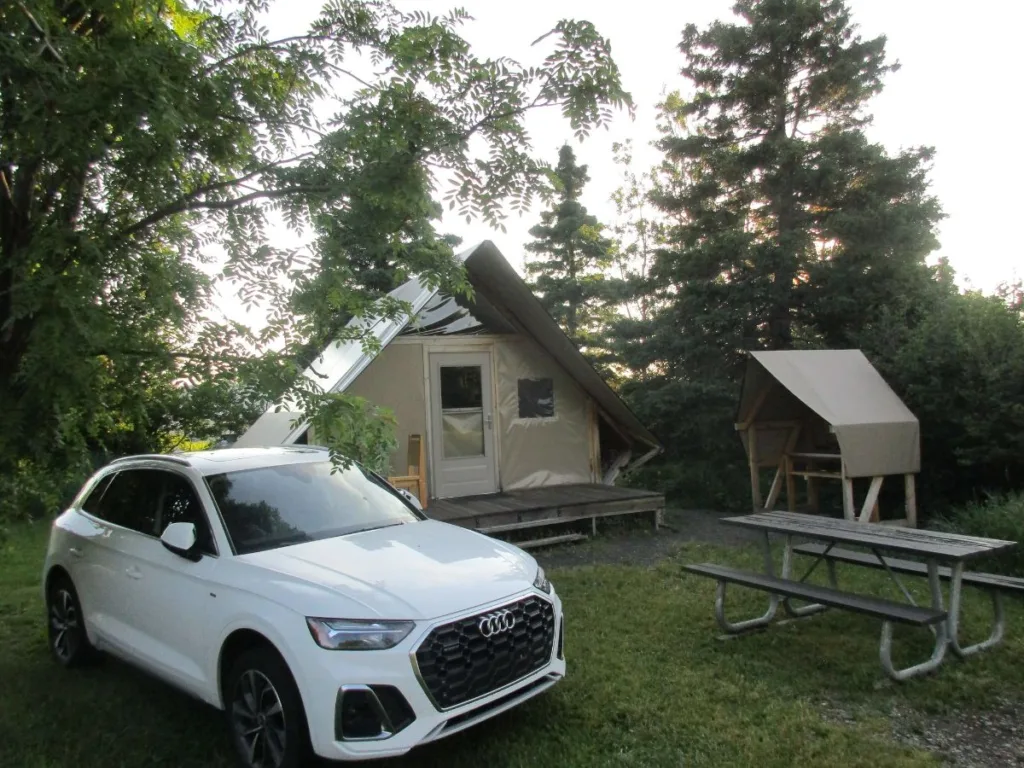 A cozy oTENTik tent in Forillon National Park, featuring a canvas and wooden frame with a small porch, surrounded by trees. A white Audi SUV is parked nearby, and a picnic table with a covered bench is situated in the grassy area, creating a picturesque camping setup.